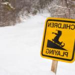 一个明黄色的标志从雪中伸出来. 它有一个孩子在雪橇上的图片，上面写着“孩子们在玩耍”."